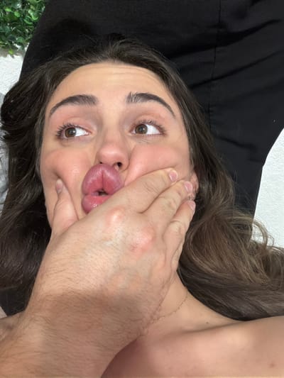 The image presents a close-up, angled view of an adult female's head and upper torso, observed from a slightly elevated perspective. The female, possessing long, dark brown hair and brown eyes, directs her gaze upwards and to the right of the frame. Her facial expression is characterized by significantly puckered lips, forming an exaggerated 'fish face' or 'duck face,' with her cheeks visibly indented. A thin gold chain necklace is visible around her neck. An adult hand, exhibiting visible hair on its dorsal surface, is positioned over the lower left side of the female's face, applying inward pressure to her cheek and chin area, which contributes to the facial distortion. The background features an indistinct arrangement of green foliage in the upper left, contrasting with a lighter wall. To the upper right and center, a dark, black fabric element, possibly clothing or bedding, is visible against a lighter wall section. The scene is located in East Naples, United States of America.