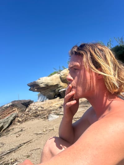 A fair-skinned male, positioned in profile facing right, is depicted on a sandy beach. He possesses long, wavy blonde hair and a light mustache. He is shirtless, revealing a tanned complexion, with vertical shadows from an unseen source falling across his face and neck. The male holds a white cigarette between his right index finger and thumb, with the cigarette positioned at his mouth as if actively smoking. The foreground consists of light brown sand interspersed with small debris. In the midground and background, large, weathered light-colored rock formations are visible, one featuring a distinct, eroded shape. Sparse green vegetation grows on and behind these rocks. The sky above is clear and bright blue. The scene is illuminated by strong natural sunlight, casting distinct shadows. The described location is Coalcliff, Australia.