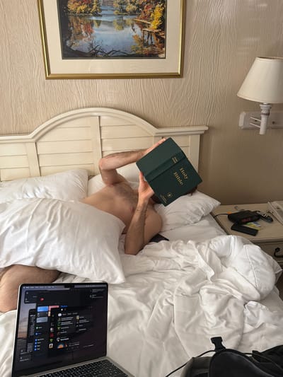 A person lies in a bed, reading a green Holy Bible held above their face. A laptop sits open on the bed next to them. The bed has white bedding and a white headboard. A painting hangs on the wall behind the headboard, and a lamp is mounted above the painting. A bedside table with various items is visible to the right of the bed. The location is implied to be Laconia, United States of America.