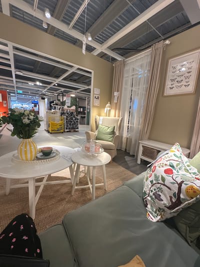 The image depicts an indoor scene, likely a furniture store showroom. In the foreground, a pale green sofa is visible, adorned with a patterned pillow featuring birds and fruit. To the left of the sofa, a round white coffee table sits on a woven rug, with a yellow and white striped vase filled with white flowers, a bowl, and a small plate on top. A smaller white side table is positioned to the right of the coffee table, holding a glass dome with a pink item inside. Further back, a cream-colored armchair with a green cushion is visible, next to sheer white curtains and beige drapes. A framed artwork of butterflies hangs on a beige wall. Behind the furniture display, rows of shelves stocked with various items, including baskets and what appear to be textiles, are visible, suggesting a retail environment. The ceiling features exposed metal beams and lighting fixtures. The scene is well-lit and presents a domestic, furnished setting within a larger commercial space. No specific location within Siero, Spain, can be identified from the image alone.