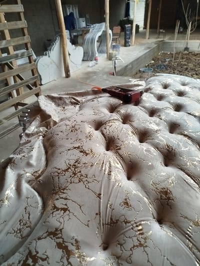A close-up shot captures a button-tufted sofa, its fabric featuring a light base with golden vein-like patterns. A red staple gun rests atop the upholstery. In the blurred background, a workshop setting includes wooden ladders, ornate white carved structures, and construction debris, indicating a furniture-making or renovation environment. The scene focuses on the upholstery process, suggesting a work-in-progress.