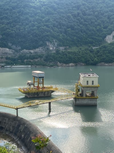 This is an eye-level, outdoor, medium shot of the Zhinvali Dam in Zhinvali, Georgia. The dam is a concrete arch dam and spans the Aragvi River. In the background, a lush, green forest covers the mountainside. The foreground features the dam itself, with a curved concrete structure dominating the lower portion of the frame. The water in the reservoir is a muted green and reflects the sunlight, creating a shimmering effect. Two yellow, metal structures rise from the water, connected by a walkway. The structure on the right has a white, two-story building on top, with windows and a small flag. The structure on the left has a taller, grey-topped building supported by yellow beams. The scene is serene and shows the impressive scale of the dam and its integration into the natural landscape.
