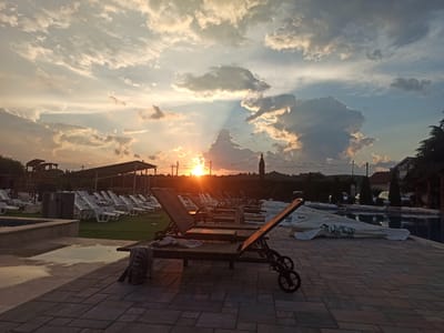 The image depicts an outdoor swimming pool area at sunset in the Sopot Urban Municipality, Serbia. In the foreground, several lounge chairs are arranged on a paved patio. Some of the lounge chairs have towels draped over them. Beyond the lounge chairs, there is a lawn with rows of white lounge chairs. A swimming pool is visible to the right, with some umbrellas covered and stacked nearby. In the background, there are buildings, trees, and a silhouette of a small tower or structure. The sky is filled with dramatic clouds illuminated by the setting sun, casting a warm glow over the scene and creating a serene atmosphere.