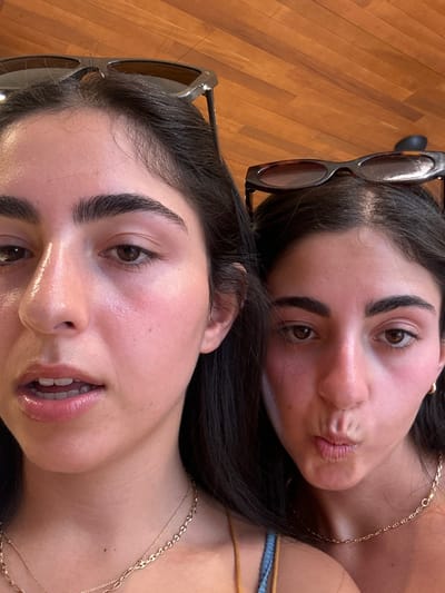The image displays a close-up, head-and-shoulder view of two young adult females. Both individuals have dark hair, dark eyebrows, and are observed with a fair complexion exhibiting minor redness on their cheeks and noses. The female positioned in the foreground on the left has her lips slightly parted, revealing her upper teeth. She wears a thin gold chain necklace. A pair of sunglasses with light-colored frames is perched on top of her head. The female on the right has her lips pursed in a 'kissing' or 'pouting' expression. She wears a thin gold chain necklace and a small gold hoop earring on her visible right ear. A pair of dark, possibly tortoiseshell-patterned, sunglasses rests on top of her head. The background consists of a uniform surface of light brown wooden planks with visible grain, indicative of an indoor setting. The image contains no visual identifiers to confirm the location in the city of San Miguel de Cozumel, Mexico.