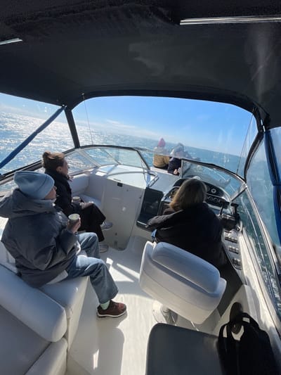 The image depicts the interior of a white motorboat underway on a sunlit body of water, presumably near Chereshnya, Russia, under a clear blue sky. Five individuals are present within the boat. In the foreground, a person wearing a light blue beanie, a dark grey puffer jacket, and light blue jeans sits on a white padded bench, holding a white disposable cup. To their right, another individual with dark hair, wearing a dark jacket, is also seated on a white bench, holding a similar white disposable cup. Further right, at the helm, a person with brown hair, dressed in a dark jacket, operates the boat, facing forward towards the windshield and open water. In the bow section, two additional individuals are visible from behind, facing forward; one wears a light-colored beanie and a tan jacket, and the other wears a dark jacket. The water surface shimmers with reflected sunlight. A black soft-top canopy extends overhead, covering part of the cockpit. The boat's helm features a steering wheel and various control panels. A black bag is visible on a seat to the far right.