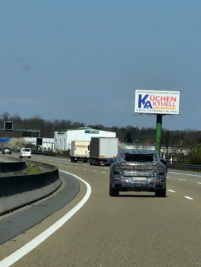 This image captures a daytime scene on a multi-lane German Autobahn, likely the A3, in the vicinity of Seligenstadt, Germany, given the billboard's reference to nearby Hanau and Weiterstadt.

In the foreground, a heavily camouflaged dark-colored SUV, identifiable as a prototype or test vehicle by its patterned wrap, occupies the right lane. Its German license plate reads "M CI 4734". Directly ahead of it in the same lane is a white semi-trailer truck. Another white semi-trailer truck is visible further ahead in the adjacent left lane, followed by a white van and a dark passenger car. The road surface is asphalt with distinct white lane markings, and a concrete barrier forms a curving median to the left.

On the right side of the highway, a large billboard stands on a green pole. The billboard advertises "KÜCHEN AKTUELL" (Kitchens Current), identifying itself as "DEUTSCHLANDS KÜCHENMARKTFÜHRER" (Germany's kitchen market leader) with additional text listing locations "IN BRÜHL, WEITERSTADT UND HANAU". Behind the billboard and trucks, commercial or industrial-type buildings with flat roofs are visible, interspersed with trees. An overhead gantry, possibly for electronic signage, spans the highway further back on the left. The sky is clear and light blue, and the overall lighting suggests midday conditions.