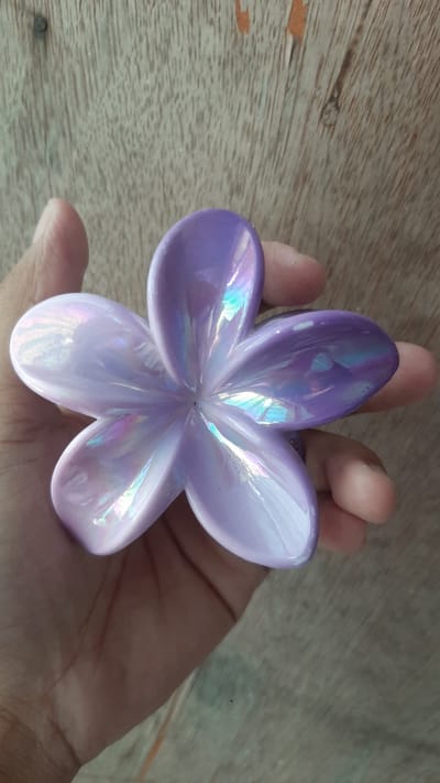 A close-up, overhead shot captures a hand holding a five-petal, iridescent purple flower ornament against a weathered wooden background. The ornament exhibits a pearlescent sheen, with highlights reflecting various colors. The wooden surface appears to be a table or floor, with visible grain and some wear marks. The image is taken indoors, likely in a domestic setting. No specific landmarks are identifiable to pinpoint the location within Kawit, Philippines.