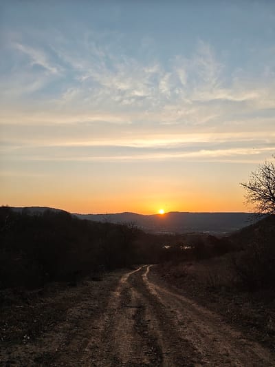 The image displays a rural landscape near Opaka, Bulgaria, at either sunrise or sunset. A winding dirt path, exhibiting tire tracks and uneven texture, extends from the foreground into the midground, subtly curving to the viewer's left. The immediate ground cover is sparse, consisting of dry earth and minimal vegetation. Dark, silhouetted hills and scattered, leafless deciduous trees and shrubs flank the path and recede into the distance. In the far background, a brilliant orange-yellow sun disc is partially visible on the horizon, casting a warm glow across the sky, which transitions from deep orange at the horizon to pale yellow and light blue, dotted with wispy white clouds at the zenith. Below the sun, a cluster of dark structures, indicative of a town (presumably Opaka), is visible in a valley. A small body of water reflects light to the right of the path in the mid-distance. No human figures or animals are present.