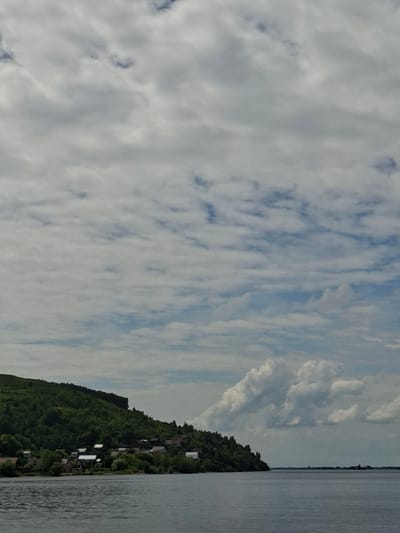 The image depicts a wide body of water in the foreground, likely a river, with gentle ripples on its surface. In the mid-ground, a densely forested hillside slopes down to the water's edge. A small village or settlement is nestled at the base of the hill, characterized by numerous houses with visible roofs. The vegetation is lush and green, covering the entire hillside. The sky above is filled with a mix of clouds; some are thin and wispy, creating a dappled effect, while others are larger and more voluminous, appearing as puffy white formations. The overall scene suggests a tranquil, rural landscape. This scene is consistent with the appearance of the Volga River near Verkhny Uslon, Russia, a region known for its scenic riverbanks and forested hills.