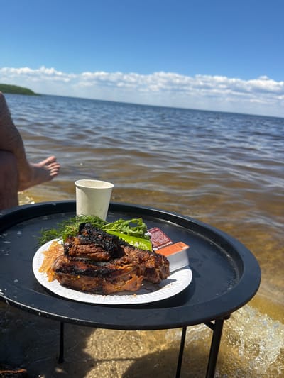 A table is set up on the shore of a body of water, with a plate of food and a cup on it. The food appears to be grilled meat with some greens and a sauce. A person's legs and feet are visible in the water to the left of the table. The water is calm with small waves lapping at the shore. The sky is blue with some scattered clouds. The scene is likely Lebedivka, Ukraine, given the context.