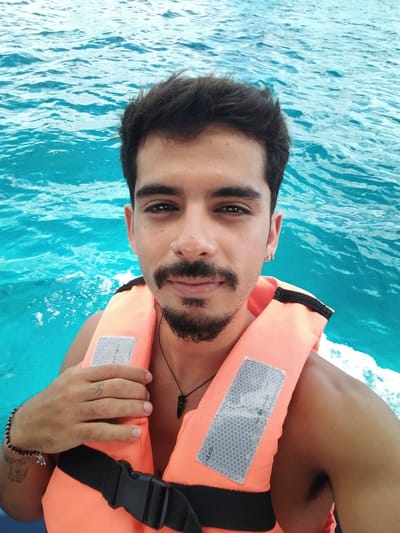 A man wearing a life jacket is on a boat. He has short dark hair, a mustache, and a beard. He has a nose ring and an earring. He is wearing a necklace. He has a tattoo on his left hand and a bracelet on his left wrist. The water is turquoise. The location is likely near San Miguel de Cozumel, Mexico, based on the color of the water and the man's appearance.