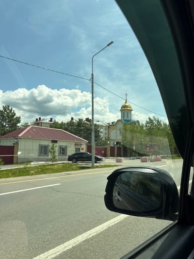 A car's side mirror reflects the interior of the vehicle and its passenger, with a faint image of a person driving. The car is on a paved road with yellow and white lane markings, suggesting it is in an urban or suburban area. To the left of the road, a low, rectangular building with a red roof and beige walls is visible, bordered by a red fence. Further down the road, a building with a golden dome and blue accents, resembling a church, stands amidst green trees. The sky is blue with scattered white clouds. The scene appears to be in the city of Sosnovka, Russia, based on the surrounding architecture and vegetation.
