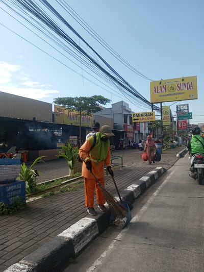 The image depicts a street scene in an urban area. A street sweeper, wearing an orange uniform, a green vest, and a mask, is using a broom to sweep debris into a dustpan. A woman in a hijab is walking along the sidewalk, carrying two bags. A motorcyclist wearing a green jacket is driving down the street. Above the sidewalk, there are numerous overhead power lines, and signage for the "Alam Sunda" restaurant. The signs indicate it's a place to eat and there is parking available. The location, based on the street signs, is near Term Cipanas and Istana Cipanas, implying it may be in or near Cipanas. Based on text on a nearby wall it is probably in Cipanas.