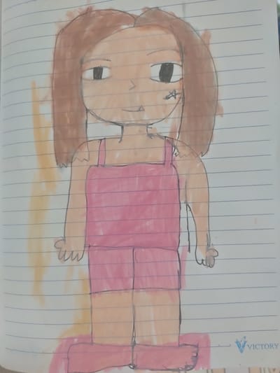 A child's drawing depicts a stylized figure with brown hair, light skin, and black eyes. The figure wears a pink outfit, possibly overalls or a romper, and pink footwear. A faint yellow or orange wash is present on the left side of the drawing. The drawing is on lined paper, suggesting it is a page from a notebook. In the bottom right corner, a logo reading "VICTORY" is visible, with a stylized blue 'V' emblem. The drawing is not located in San Pascual, Philippines.