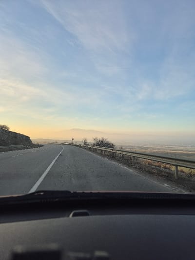 The image presents a forward view from the interior of a vehicle on a paved two-lane roadway in Bogyovtsi, Bulgaria. The asphalt road, marked with a solid white line on its left edge, curves gently to the right in the mid-distance. To the immediate left, a rocky embankment ascends, paralleled by a metallic guardrail. To the right of the road, a similar metallic guardrail borders a steep decline into an expansive, flat agricultural landscape that extends to a hazy mountain range on the distant horizon. The sky is predominantly light blue with sparse, wispy cirrus clouds, transitioning to a soft yellowish-orange hue near the horizon, indicative of either sunrise or sunset. The lower foreground of the image displays the dark grey dashboard and the black bases of windshield wipers belonging to the observation vehicle. No individuals or specific actions are discernible within the frame beyond the implied presence of a vehicle operator.