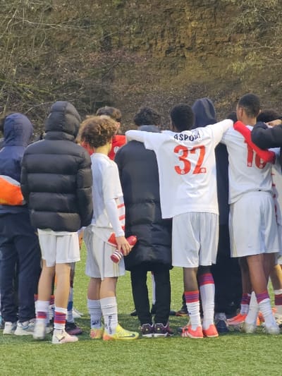 A group of young men or boys, likely a soccer team, are standing in a line outdoors on what appears to be a sports field. The image is taken from slightly behind and to the side of the group, showing most of them from the back.
The setting consists of a bright green artificial turf field at the bottom right of the image, where the individuals are standing. Behind them, to the left, is a natural, earthy embankment or hillside covered with sparse, brownish-green vegetation, suggesting a wooded or uncultivated area. The lighting suggests it is daytime, possibly overcast, as there are no strong shadows.
The individuals are dressed in sports attire. Most are wearing white short-sleeved jerseys with white shorts and long athletic socks that extend up their calves. Various colorful soccer cleats are visible on their feet. Some individuals are wearing additional outerwear: one person in the middle wears a black puffer jacket over their uniform, and another towards the left is wearing a dark blue hooded jacket. One person, closer to the front of the line, is holding a red water bottle with white stripes.
On the back of one of the white jerseys, visible in the center of the image, the partial text "4SPOR" and a large red number "37" are clearly printed. Other details like hair textures are visible, with some having curly hair and others shorter styles. The overall activity seems to be a team gathering, possibly before or after a game or practice session, as they are lined up and facing forward, suggesting they are attending to a speaker or looking in a particular direction.