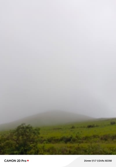 An outdoor medium shot captures a misty, rolling landscape with green grass and a small tree in the foreground. A distant hill is obscured by fog. The bottom of the image displays text and a red dot, indicating it's a photograph taken with a CAMON 20 Pro. The camera settings are also listed: 24mm f/1.7 1/249s ISO50. The location is identified as Nguroje, Nigeria.