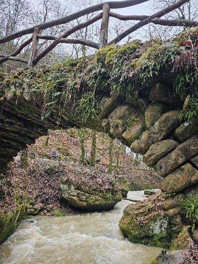 The image displays a rustic stone arch bridge, heavily overgrown with green moss and various ferns, located within the natural landscape of Waldbillig, Luxembourg. A weathered wooden railing, constructed from uneven logs and branches, spans the top of the bridge. Beneath the bridge, a turbulent, light brown river flows rapidly through a narrow gorge, exhibiting white water and foamy rapids. The riverbanks are characterized by large, irregular boulders, also covered in moss, and a dense layer of fallen reddish-brown leaves. In the background, numerous bare deciduous trees stand against an overcast, light grey sky. No individuals or animals are visible.