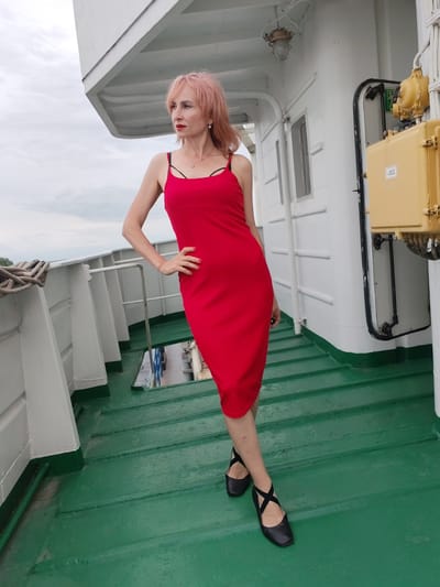 A woman with pink hair stands on the green deck of a white ship, looking to her left. She is wearing a form-fitting red spaghetti strap dress and black ballet flats with criss-cross straps. Her left hand is on her hip. The ship's deck is lined with white railings, and a white superstructure rises behind her. To her right, a yellow electrical box with some visible text is mounted on the ship's wall. The sky is overcast, suggesting cloudy weather. The scene appears to be daytime. The location context is Bereg, Russia.