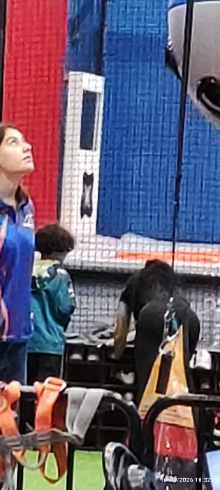 A woman in a blue shirt looks up at a black camera mounted on a black pole. Behind the woman, there is a blue, red, and white wall with a black stripe in the middle of the white section. To the right, a messy collection of items includes harnesses, bags, and other gear. A person wearing a green shirt stands to the side.