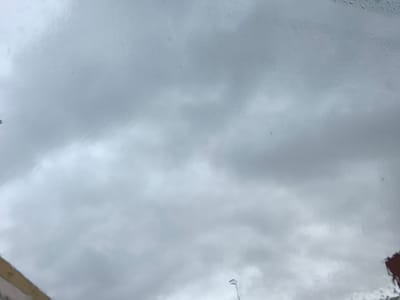 The image displays an overcast sky viewed through a wet surface, likely a window or windshield, suggesting rainy weather. The sky is a uniform grey, indicating heavy cloud cover, with subtle variations in tone reflecting cloud formations that are largely obscured and distorted by water on the glass.
Numerous raindrops and streaks of water are visible across the transparent surface, particularly dense in the upper right and central regions, blurring the view beyond. In the bottom left corner, a partial view of a light-colored building or structure with a brownish-tan top edge is discernible. Below this edge, some dark, indistinct shapes are present, and faint, illegible marks, possibly text, are barely visible. Near the bottom center, a slim, light-colored object, potentially the top of a streetlight or pole, points upwards, also distorted by the wet glass. The bottom right corner shows a fragment of a reddish-brown structure, possibly a tiled roof.
The scene depicts a dull, cloudy, and rainy day. No people or specific activities are visible. The elements suggest an urban or semi-urban setting in Nàquera / Náquera, Spain. The time of day appears to be daytime, despite the heavy cloud cover. No clearly readable text is present.