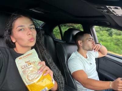 In this media file, two individuals, a man and a woman, are inside a car. The man, on the left, is wearing a white t-shirt and a bracelet on his left wrist. He has short hair and a beard and appears to be looking down, with his hand near his face. The woman, on the right, is holding a yellow bag of oats. She has long, dark braided hair and is wearing a black top. Her nails are styled with a pink and white French tip design with rhinestones. She is looking towards the camera with pursed lips.

The scene is set inside a car, with the interior visible, including car seats and dashboard elements. Outside the car, a blurred green landscape suggests the vehicle is in motion, likely on a road surrounded by nature. The lighting suggests it is daytime. The overall mood appears casual and relaxed.

Visible text on the yellow bag includes "500g", "OBECЕН", "ИНФИ", and "DR. RESKIN". The text is primarily in a Cyrillic script. The location context provided is Хаджийско, Bulgaria.