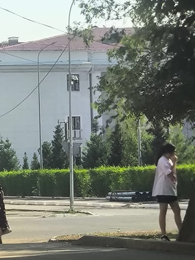 A daytime, medium-shot image captures a street scene in Abay, Kazakhstan. In the foreground, a person with dark hair, wearing a white t-shirt and black shorts, stands on a sidewalk next to a tree trunk. They are partially obscured by the tree and appear to be looking down or to their right, possibly touching their face with their hand. Their stance suggests a relaxed or contemplative mood.
Behind the person, a row of manicured green bushes forms a border along a paved area. In the middle ground, a building with a tiled red roof and white walls is visible. The building has several windows, one of which is partially obscured by a street light pole. The architecture suggests a formal or institutional building.
Further back, a line of dark green trees and foliage adds depth to the scene. The sky is a pale blue with some overcast. On the paved area behind the bushes, a few cylindrical objects are stacked, possibly construction materials or debris. A street light pole stands prominently in the middle of the frame, with wires extending from it. The presence of the building, the manicured bushes, and the street lights suggest an urban or semi-urban environment. The time of day appears to be sunny, likely late morning or afternoon, given the brightness and the shadows. There is no visible text in the image.