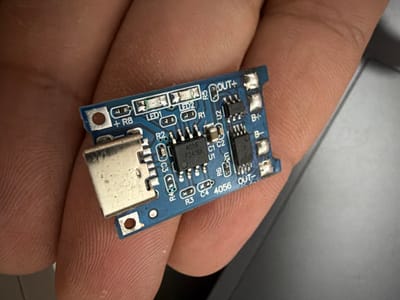 The image shows a close-up of a person's hand holding a small blue circuit board. The circuit board has various electronic components, including a USB-C connector, resistors labeled R1, R2, R3, R4, R5, LEDs labeled LED1 and LED2, capacitors C1, C2, C3, and C4, and an integrated circuit labeled U1 with the markings "4056 F2418X". There are also connection points labeled "+R8", "OUT+", "B+", "B-", and "OUT-". The hand's skin texture and fingerprints are visible. The background is blurry and appears to be a gray, flat surface. There is no indication of the location in Torremolinos, Spain.