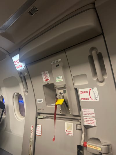 A close-up shot depicts an airplane emergency exit door and its surrounding paneling. The door features instructions in English and Russian, including "Open," and "Pull cover to open." Red and yellow safety components are prominently displayed, along with additional instructions for emergency opening. A lower panel includes an illuminated "EXIT" sign written in both English and Russian. The overall color scheme is grey and white, with black text and red accents. The lighting creates shadows that highlight the various features of the door and paneling.