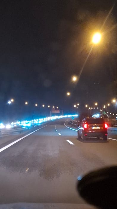 A night road scene shows a dark car ahead with illuminated tail lights. Rows of yellow and blue streetlights line the road. The road's surface is visible, marked with white lines. A partial view of the camera-holder is on the upper right corner. The scene appears to be recorded from the dashboard camera of a vehicle traveling on the road.