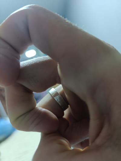 This is a close-up, vertically oriented, over-the-shoulder shot of a person's hands interlocked. A silver ring with a textured, checkered pattern is visible on the ring finger of the lower hand, which is clasped over the upper hand. The skin texture, including faint lines and slight hair on the knuckles, is discernible. The lighting suggests an indoor setting with a bright, out-of-focus light source visible in the background, creating a soft glow and casting subtle shadows on the hands. The overall impression is one of intimacy or contemplation, with the focus solely on the hands and the ring. There is no discernible information about the location in Uchquduq, Uzbekistan.
