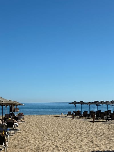 A bright, sunny day at a beach in Rethymno, Greece. The sand is light brown and textured. In the foreground, lounge chairs and umbrellas are neatly arranged on the sand. To the left, several light-colored umbrellas and lounge chairs are visible, some with towels draped over them. To the right, a row of dark umbrellas shades more lounge chairs. A few people are present; one person is sitting under an umbrella on the right side, facing the sea. Another person is visible in the water to the left, further out from the shore. The sea is a calm, deep blue, meeting a clear, bright blue sky at the horizon. The overall scene is tranquil and indicative of a typical beach day.