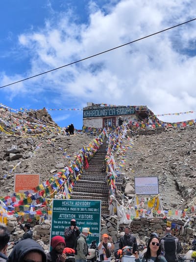 The image captures a view of the Khardung La Pass in Sasoma, India, a mountain pass known for being one of the highest motorable roads in the world. A steep set of metal stairs, adorned with numerous colorful prayer flags, ascends a rocky hillside towards a building at the summit. The building, with signage that reads "Khardung La," is also decorated with prayer flags. Several signs are visible at the base of the stairs, including one labeled "HEALTH ADVISORY" for Khardungla Pass, providing altitude information and advice for visitors. Another sign, with orange text on a white background, states "Visitors are requested to kindly avoid stepping onto the holy images..." A group of people, dressed in casual and outdoor wear, are gathered at the bottom of the stairs, some appearing to be tourists. A few individuals are seen interacting with each other, and some are holding cameras or phones. The environment is mountainous and rocky, with a clear blue sky filled with white clouds. A power line stretches across the top of the frame.