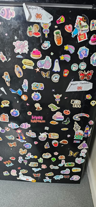 This is a close-up, top-down shot of a refrigerator door covered in a variety of stickers. The stickers depict a range of whimsical and often childlike imagery, including cartoon characters, animals, food items, and Halloween-themed illustrations. Several stickers feature bright colors and playful designs, such as unicorns, mermaids, octopuses, and smiling penguins. There are also some butterfly and flower stickers, along with a few that appear to be handmade or from a specific event, indicated by text like "happy halloween," "trick or treat," and "Cosy Blanket £8." One sticker is a white paper label with red writing that appears to say "Helpwr the" and "elpwr Heddin." The overall impression is one of personal decoration and a festive atmosphere, likely in a home setting. The refrigerator door itself is black, showing some signs of wear and smudging, and is set against a speckled grey backsplash and a white ceiling. The lighting suggests an indoor environment, possibly during the day given the clarity of the image. The stickers suggest a mix of general interests and possibly seasonal celebrations.