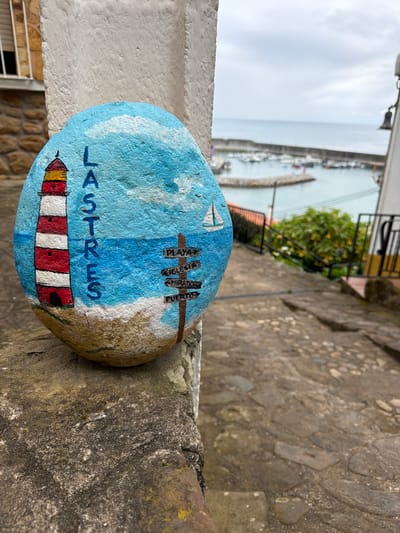 The image displays a painted rock positioned on a coarse stone ledge, overlooking the coastal village of Lastres within the municipality of Colunga, Spain. The rock's surface is painted with a scene depicting a blue sky with white clouds, a red and white striped lighthouse on a sandy beach, and a blue sea featuring a white sailboat. The word "LASTRES" is painted vertically in blue on the rock, alongside a brown painted signpost with four directional indicators labeled in Spanish: "PLAYA" (beach), "IGLESIA" (church), "MIRADOR" (viewpoint), and "PUERTO" (port).

The stone ledge is part of a descending, irregularly paved stone path, which recedes into the background. A black metal railing runs along the right side of this path. In the background, a harbor is visible, containing numerous boats moored behind a concrete breakwater. The open sea extends to the horizon under an overcast sky. Architectural elements include a building facade with a visible window and a rolled-up blind on the left, and a partial view of a white building with a dark bell on the right. Green foliage is present behind the right-side railing. No individuals are depicted in the scene.