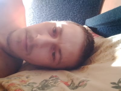 A young Caucasian male lies on his back on a bed with a patterned floral comforter. The man has short brown hair, a light complexion, and a light beard. The room appears to be dimly lit by sunlight streaming in from the left side of the frame, casting shadows and highlights across the man's face and the surrounding environment. The man is positioned at an angle, with his head closer to the camera and his body extending away from it. The background shows a dark, textured surface, possibly a wall or furniture, and a blue, fuzzy material, which could be a blanket or upholstery. The overall impression is of a candid, personal moment, possibly taken shortly after waking up or while resting. There is no indication of the location being in the city of Ушья, Russia.