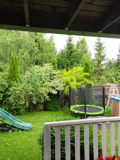 The image shows a backyard in Ермолино-2 Снт, Russia. The scene is dominated by lush green grass and a variety of trees and bushes. In the foreground, a wooden railing is visible, suggesting a porch or deck. To the left, a green children's slide is positioned over a green lawnmower. Further into the yard, a black trampoline with a neon green mat and enclosure is set up. Behind the trampoline, a fence and part of a swimming pool structure are visible. The overall environment is that of a well-maintained suburban backyard, likely used for recreation. The sky is overcast, indicating a cloudy day.