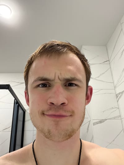 An eye-level indoor photograph depicts an adult male from the chest up, positioned centrally. He has wet, light brown hair and a light skin tone. His face displays a quizzical or slightly confused expression, characterized by furrowed eyebrows creating horizontal wrinkles on his forehead and between his brows, while his mouth forms a subtle, closed smirk. Small red marks are visible on the left side of his forehead and across the bridge of his nose. He is shirtless, with visible water droplets on his left shoulder, and wears a black braided necklace. The background features light-colored walls, appearing to be white or light grey ceramic tiles with subtle dark grey veining, consistent with a modern bathroom. A dark-framed shower enclosure is partially visible on the left. A circular overhead light fixture emits bright illumination from the top left of the frame. The specific location in the city of Tliustenkhabl', Russia, cannot be determined from the image.