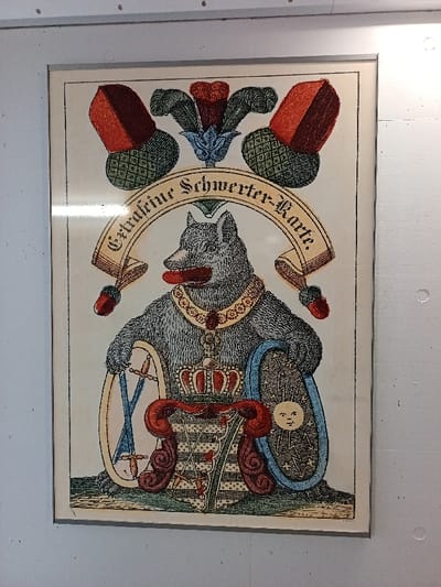 A framed, hand-colored illustration is centrally mounted on a white, possibly wooden, surface with three visible silver fasteners along the top edge, in a location within Altenburg, Germany. The print, titled "Extrafeine Schwerter-Karte" in German on a curved banner above the main subject, depicts a standing, anthropomorphic gray wolf-like creature with a protruding red tongue. The wolf wears a golden and red jeweled collar. Its right paw holds a circular shield adorned with crossed blue swords and two small red objects, while its left paw secures an oval shield featuring a stylized human-like sun or moon face with radiating lines and stars on a dark, patterned background. A red crown rests upon a large heraldic shield positioned below the wolf's torso, displaying geometric patterns and small red heart-like symbols. Above the banner, two red and green acorn-like motifs flank a central blue and red feathered plume. The entire illustration is contained within a thin black border and an off-white mat, protected by glass within a light gray frame.
