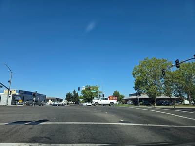 The image depicts a street-level view of an intersection in West Sacramento, California, USA, on a clear, sunny day. The intersection features multiple lanes of asphalt road with white lane markings and a stop line visible in the foreground. Traffic lights are present at the intersection, with a sign indicating "Harbor Blvd".
On the left side of the intersection, a commercial building with blue and gray signage is visible, identifying it as "E-MOG" and "Auto Repair & Service". A dark-colored pickup truck is parked in front of the building. Further down the street, more commercial buildings and trees are seen.
On the right side of the intersection, a white pickup truck with a prominent "Carl's Jr." sign on its bed is stopped at the intersection, facing forward. Behind the pickup truck, a Carl's Jr. restaurant with a red roof is visible, with several cars parked in its lot. Large trees with green foliage are situated in front of the restaurant.
In the distance, more trees and buildings line the street, under a clear blue sky with a faint, wispy cloud. The overall scene is consistent with a suburban commercial area. There are no people clearly visible in the image.