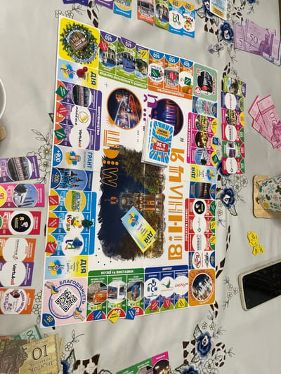 A board game, laid out on a tablecloth patterned with white, brown, and blue floral designs, is the focal point. The game board features places and organizations within the city of Vinnytsia, Ukraine and contains text in Ukrainian. Scattered around the board are stacks of play money, dice, and property cards. A few small yellow cards, presumably tokens or markers, are placed near a black cell phone. A mug with a snowflake pattern sits on a bamboo coaster in the upper center of the frame.