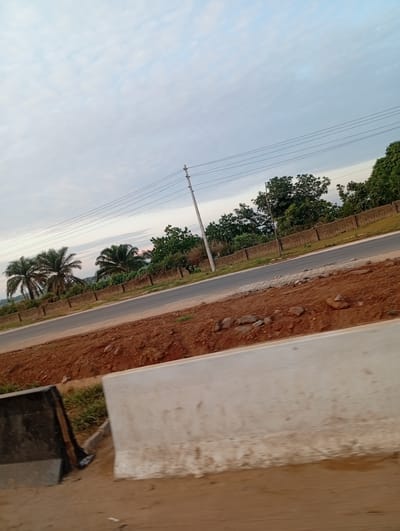 The image captures a roadside scene in Uke, Nigeria, under a cloudy, light blue and grey sky. In the foreground, a light-colored concrete road barrier is partially visible on the right, with reddish-brown soil and small scattered rocks immediately in front of it. To the left of this, a darker concrete barrier is present, with some green grass patches between it and the main dirt area. A paved grey road runs diagonally through the midground, receding from the lower left towards the upper right. Parallel to the far side of the road, a continuous low concrete block wall extends horizontally across the frame. Behind this wall, dense green foliage and trees are visible, including several prominent palm trees on the left side. A light-colored utility pole stands on the far side of the road, positioned centrally to the right, supporting multiple horizontal power lines that stretch across the sky. No individuals, animals, or vehicles are present.