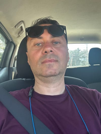 In Berbuki, Georgia, a man wearing sunglasses and a burgundy t-shirt is pictured inside a car. He is seated in the driver's seat, as indicated by the seatbelt across his chest and the car's interior visible around him. The man is looking towards the camera, taking a selfie. The background shows trees and power lines through the car's windows, suggesting an outdoor, possibly semi-rural setting. The car's interior features a headrest and a grab handle, typical for a vehicle. The man has short, dark hair and light-colored eyes. A blue cord, likely from earbuds, is visible around his neck.