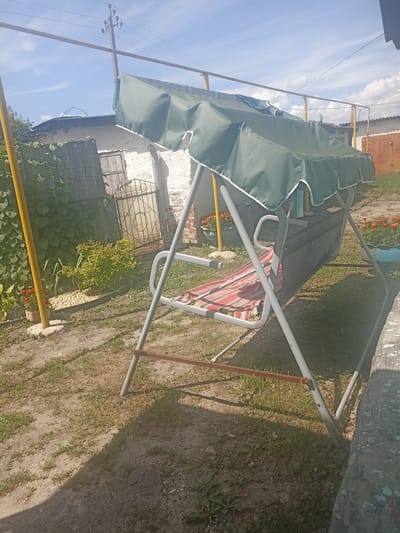 The image depicts a backyard scene in the city of Pereleshinsky, Russia, on a sunny day with scattered clouds. A green-canopied garden swing, with a striped cushion, is the central object. The swing is positioned on a dirt and grass ground, with sparse vegetation and a weathered fence in the background. To the left, a yellow pole supports a wire structure, and a dense green vine covers part of the fence. Orange flowers in pots are visible near the swing. The overall impression is one of a rural or suburban dwelling.