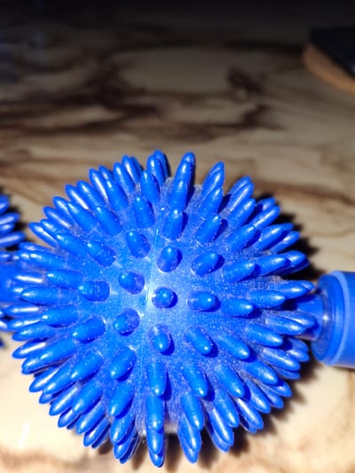 A close-up, low-angle shot focuses on a bright blue, spiky massage ball resting on a patterned surface, likely a table with a wood-grain effect. The ball's surface is covered in numerous pointed protrusions, some of which appear slightly dulled or worn. A portion of another, similar blue object is visible to the left of the main subject. The background is blurred, with the top portion of the image appearing dark and out of focus, contrasting with the well-lit foreground. The overall impression is that of a product photograph or a detail shot of an object for personal use. The location in Åsgårdstrand, Norway is not determinable from the image itself.