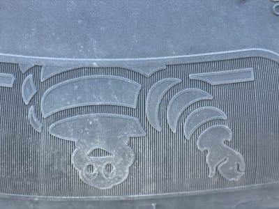 This is a close-up, high-angle shot of a dark gray rubber mat with a textured surface. The mat features a raised relief design that appears to be a stylized depiction of a pig's face on the left and a stylized bird or similar creature on the right, separated by decorative elements resembling leaves or flames. The mat is oriented horizontally, filling the entire frame. The texture of the rubber consists of fine parallel vertical lines, with the raised design elements having a slightly smoother, more worn appearance. The lighting is diffused, casting no harsh shadows. There are no people or signs of human activity visible in the image. The location in Zarech'e, Russia is not discernable from the visual information provided.