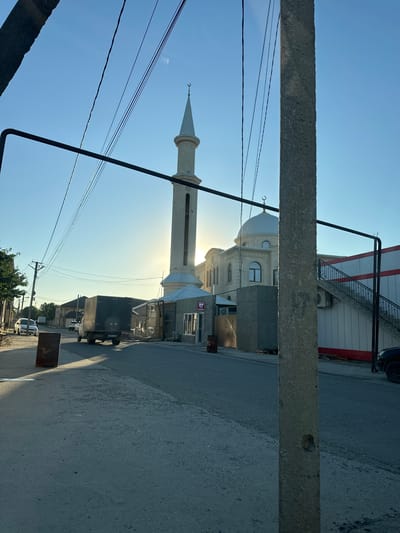 The image depicts a street scene in Miatli, Russia, on a clear, sunny day. The foreground is dominated by a rough, concrete structure, possibly a pillar or beam, running horizontally across the frame, and power lines that crisscross the sky.
In the background, a mosque with a tall, slender minaret and a dome is visible. The minaret is adorned with a crescent moon at its peak. Adjacent to the mosque, a modern building with a red trim and an external staircase with decorative railings is present. Sunlight streams intensely from behind the mosque, creating a bright glow and casting long shadows.
The street itself is paved with asphalt and appears to be lined with buildings. A dark-colored truck is parked on the right side of the road, partially obscuring a building. Further down the street, other vehicles and residential structures are visible. The overall impression is of a quiet, daytime scene in a small town or village. No people are clearly discernible, but the presence of vehicles suggests ongoing activity. No visible text is legible in the image.