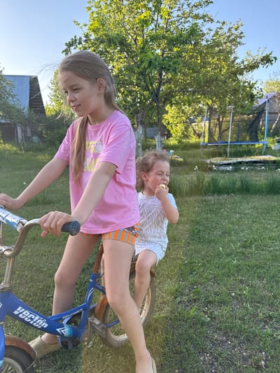 Two children are on a bicycle outdoors. One child is sitting on the handlebars of the bicycle, eating. The other child is standing and holding the handlebars. The bicycle is blue with rust and has the word "vector" on the frame. The setting appears to be a yard with grass and trees. A trampoline is visible in the background, along with a small building. The location is Галево, Russia.
