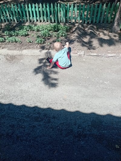 The image depicts a young child, likely a toddler, wearing a light blue shirt with red accents and red pants. The child is standing on a paved surface, possibly a walkway or road, and appears to be interacting with the ground, perhaps playing or reaching for something. A green wooden fence runs along the left side of the image, with some vegetation growing beside it. Shadows cast by what appear to be trees or foliage create dappled patterns on the ground and the child. The lighting suggests it is daytime, possibly late morning or early afternoon, given the sharp shadows. The scene appears to be outdoors, in a park or a residential area with a fence and a paved path. The overall mood is one of quiet observation and childhood play. There is no visible text in the image. The location context is Көксу, Kazakhstan.