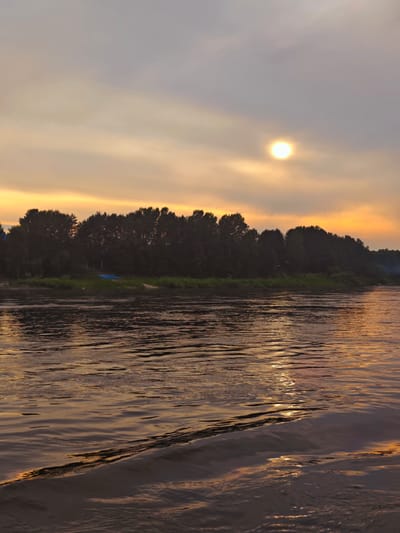 A sunset scene over a wide river, viewed from the water's surface. The sky is filled with diffused sunlight and soft clouds, tinged with yellow and orange hues, suggesting dusk. A bright, circular sun is visible in the upper right portion of the sky, partially obscured by clouds. The opposite bank of the river is lined with a dense, dark silhouette of trees. On the bank, a small, blue object, possibly a boat or cover, rests on the grassy shore. The river's surface is rippled, reflecting the colors of the sky and creating streaks of light. In the foreground, the water shows distinct wakes and wave patterns. The overall atmosphere is serene and natural, indicative of a rural or wilderness setting near YUKSEEVO, RUSSIA.